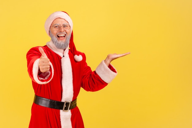 Felice uomo anziano felice con la barba grigia che indossa il costume di babbo natale che presenta lo spazio della copia per la pubblicità sul palmo, mostrando il pollice in su, consiglia. Colpo dello studio dell'interno isolato su priorità bassa gialla.