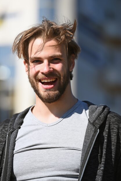 Felice uomo barbuto con acconciatura alla moda e volto sorridente in camicia soleggiata all'aperto su sfondo sfocato