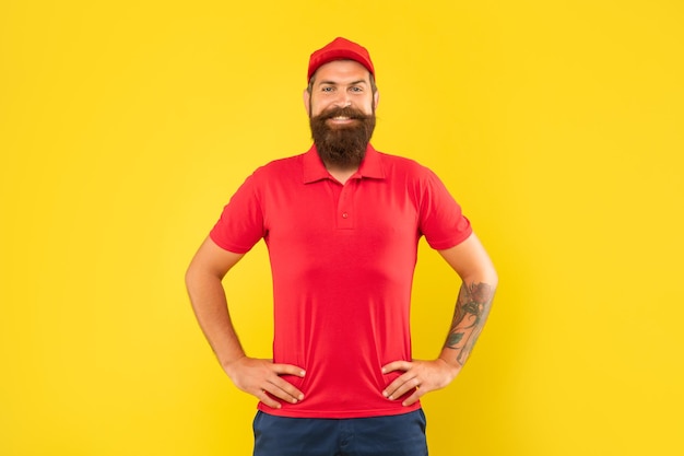 Felice uomo barbuto in berretto rosso e maglietta in piedi con braccia tatuate sui fianchi sfondo giallo casual
