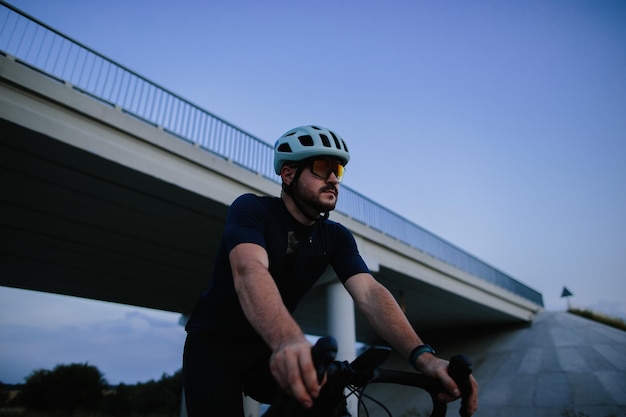 Felice uomo barbuto in casco protettivo occhiali a specchio e abbigliamento sportivo in sella a una bicicletta su una strada asfaltata fuori città la sera