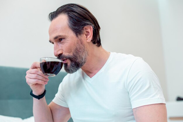 Felice uomo barbuto in maglietta bianca seduto a casa e sorridente mentre beve il caffè da una tazza di vetro