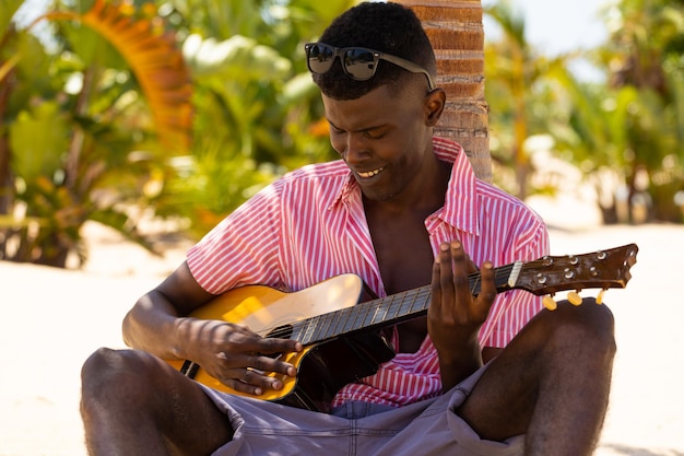 Felice uomo biraziale che suona la chitarra seduto vicino alla palma sulla spiaggia soleggiata. Musica, relax, lifestyle, estate e vacanza, inalterati.