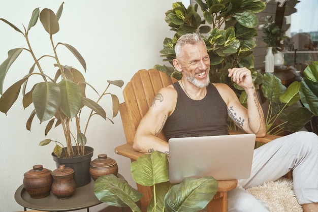 Felice uomo caucasico di mezza età in abiti casual che usa il computer portatile mentre è seduto sul balcone o