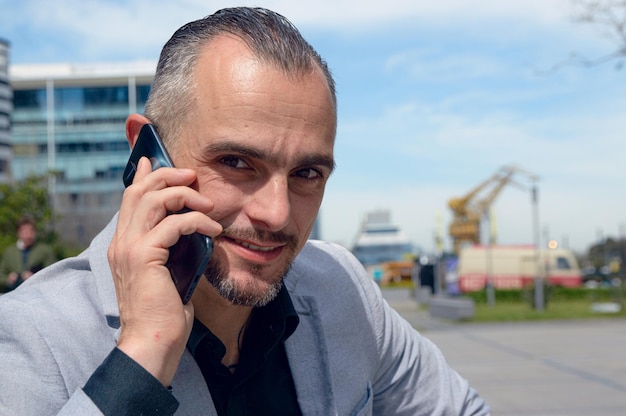 felice uomo d'affari adulto caucasico seduto sorridendo parlando al telefono all'aperto e guardando la telecamera