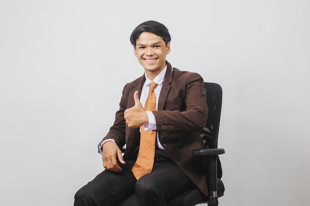 Felice uomo d'affari asiatico in giacca e cravatta è seduto su una sedia da ufficio e dà il pollice in su