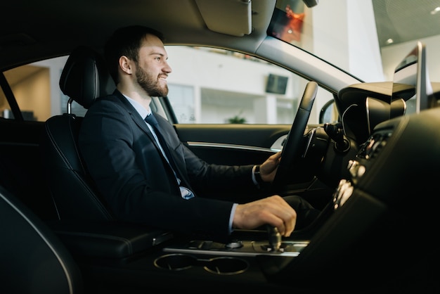 Felice uomo d'affari barbuto che indossa un tailleur è seduto al volante di una nuova auto