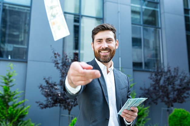 Felice uomo d'affari caucasico di successo con la barba conta, onde, lancia, mostra, ostenta sfondo di denaro contante un moderno edificio per uffici guardando la fotocamera Festeggia il successo del vincitore nelle scommesse sul mercato azionario