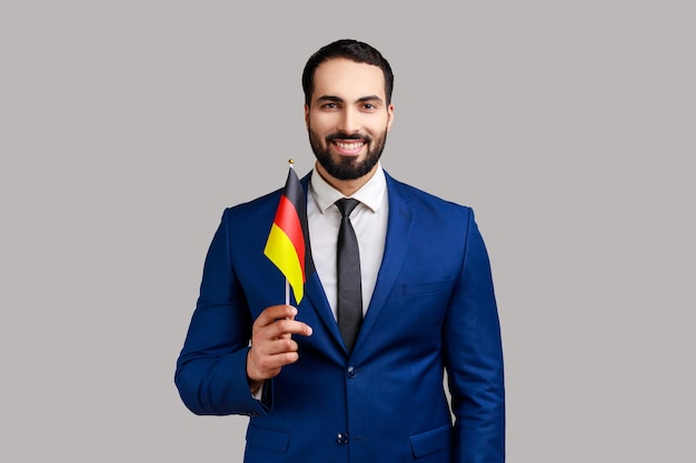 Felice uomo d'affari che tiene la bandiera della Germania che celebra il giorno della Germania il 3 ottobre esprimendo emozioni positive indossando un abito in stile ufficiale Indoor studio shot isolato su sfondo grigio