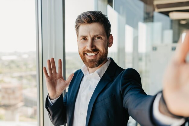 Felice uomo d'affari di successo con la barba in tuta agitando la mano alla fotocamera dello smartphone che si incontra in remoto