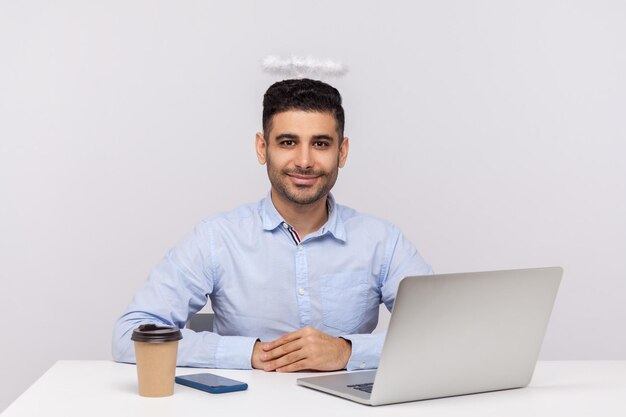 Felice uomo d'affari di successo o top manager seduto nel posto di lavoro dell'ufficio con alone di sant'angelo sulla sua testa sorridente godendo il titolo di miglior impiegato indoor studio girato isolato su sfondo bianco