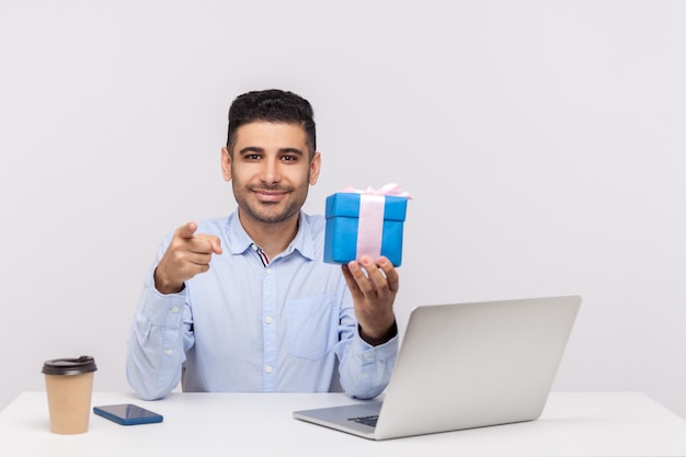 Felice uomo d'affari elegante seduto sul posto di lavoro dell'ufficio, con in mano una confezione regalo e puntando alla fotocamera, scegliendo te, mostrando il bonus presente, congratulandosi per le vacanze professionali. girato in studio isolato