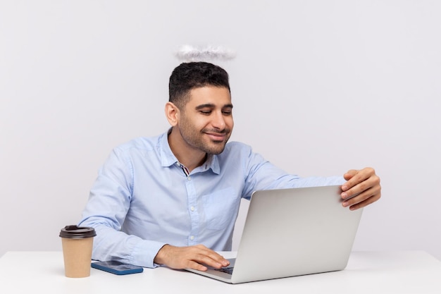 Felice uomo d'affari seduto sul posto di lavoro dell'ufficio con l'aureola di sant'angelo sulla testa il suo sorriso che esprime amore per un caro amico in videochiamata con incontri online tramite laptop studio indoor girato isolato