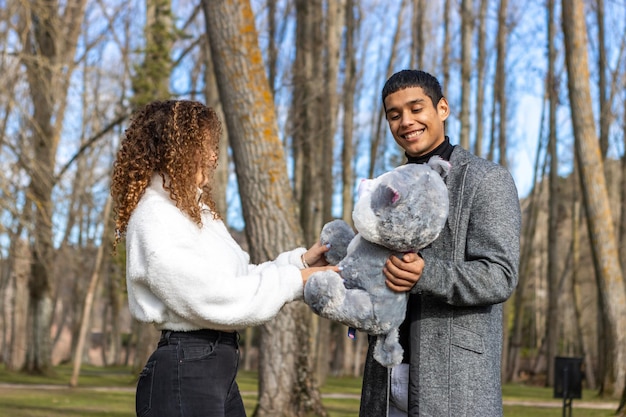 Felice uomo latino che regala un orsacchiotto alla sua ragazza Ragazzo che fa un regalo di San Valentino alla sua ragazza all'aperto