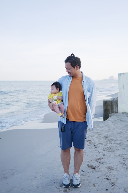 Felice vacanza in famiglia asiatica, un padre tiene il suo piccolo bambino carino in spiaggia in estate, guarda il suo bambino, viaggio in famiglia in mare