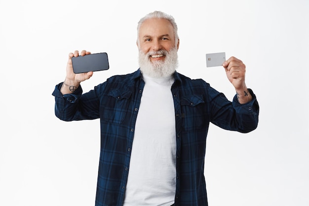 Felice vecchio barbuto che mostra lo schermo orizzontale dello smartphone e la carta di credito che consiglia l'app bancaria dell'applicazione mobile in piedi su sfondo bianco