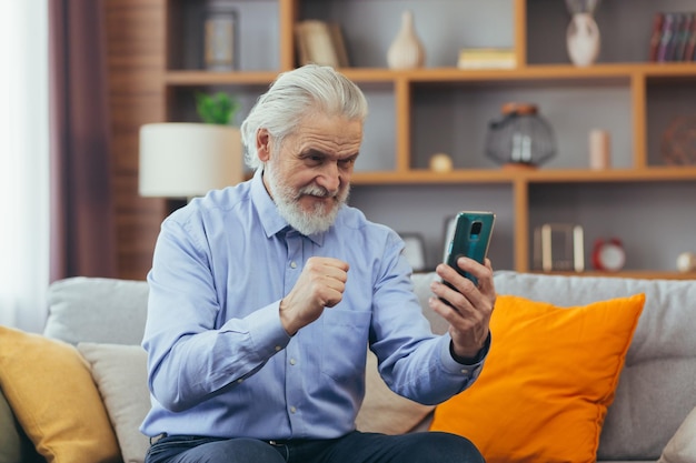 Felice vecchio usa il telefono a casa seduto sul divano utilizza applicazioni mobili