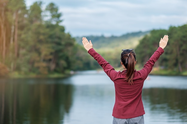 Felice viaggiatore donna vista posteriore con le braccia in alto e guardando uno sfondo di fiume e foresta