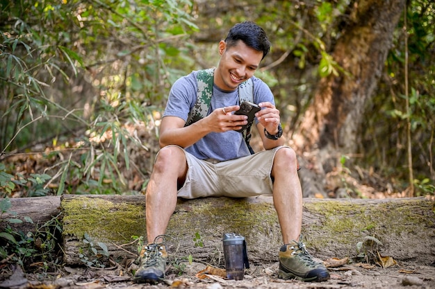Felice viaggiatore maschio asiatico o trekker che utilizza il suo smartphone mentre riposa su un tronco di legno