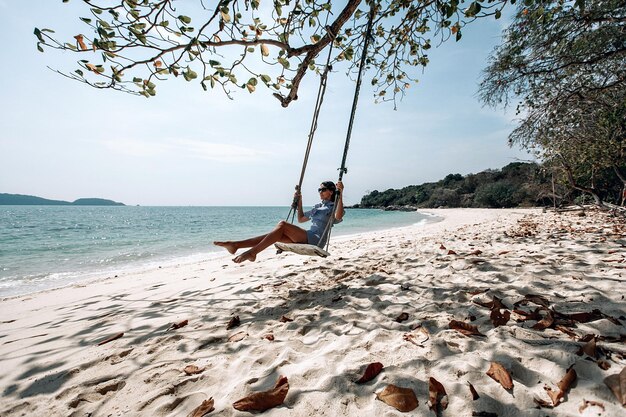 Felice viaggiatrice in abito scozzese bianco e nero e occhiali da sole neri che si rilassano sull'altalena. Spiaggia turistica del mare Tailandia, Asia, viaggio di viaggio di vacanza di vacanza estiva. Phuket. Tailandia