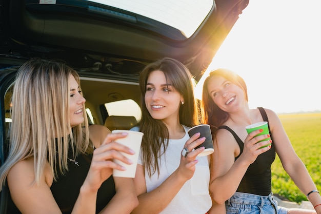 Felice viaggio su strada di tre viaggiatrici sorridenti, rilassati sul retro del veicolo con tazze di caffè da asporto alla luce del sole. Le ragazze stanno bevendo il caffè.