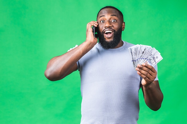 Felice vincitore! Giovane uomo afroamericano ricco in maglietta casuale che tiene banconote da un dollaro soldi e telefono cellulare con sorpresa isolato sopra la parete verde.