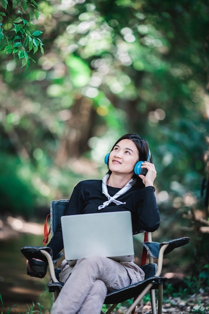 Felicemente giovane donna seduta su una sedia da campeggio che indossa le cuffie per ascoltare musica dal computer portatile mentre si rilassa in campeggio nella foresta