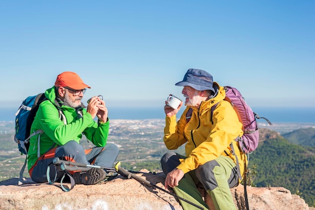 Felici escursionisti anziani con zaini che si godono una tazza di caffè facendo una pausa rilassante in cima alla collina