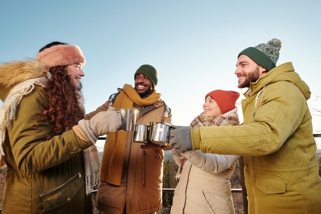 Felici giovani amici in abbigliamento invernale che si rallegrano con tè caldo e chiacchierano contro il cielo blu su montagne e foreste durante il fine settimana invernale