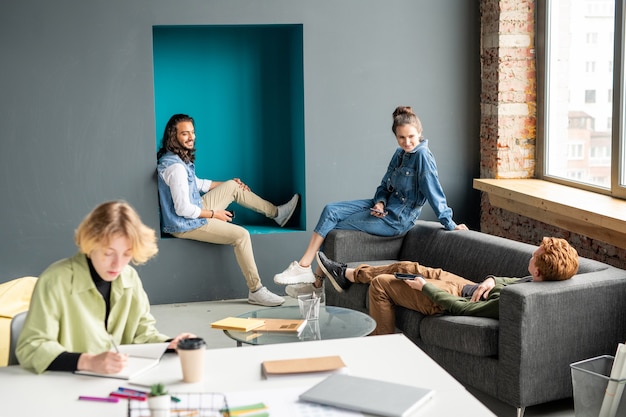 Felici giovani designer creativi in abbigliamento casual seduti vicino alla finestra in ufficio, brainstorming e discutendo i punti di lavoro alla riunione