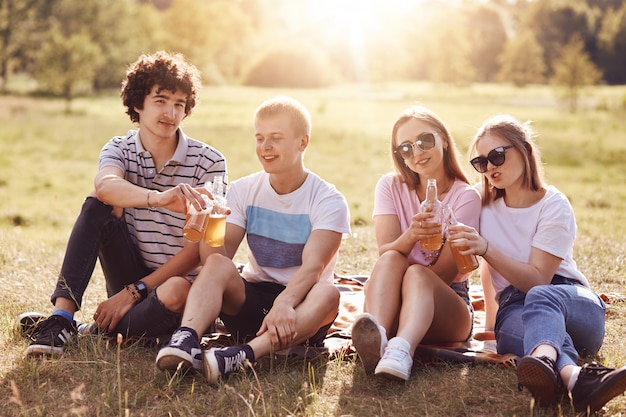 Felici ragazzi e ragazze festeggiano qualcosa, tintinnano bottiglie di birra durante il picnic, hanno espressioni positive, godono la giornata di sole estivo e la natura meravigliosa, sorridono con gioia. Amicizia e concetto di riposo