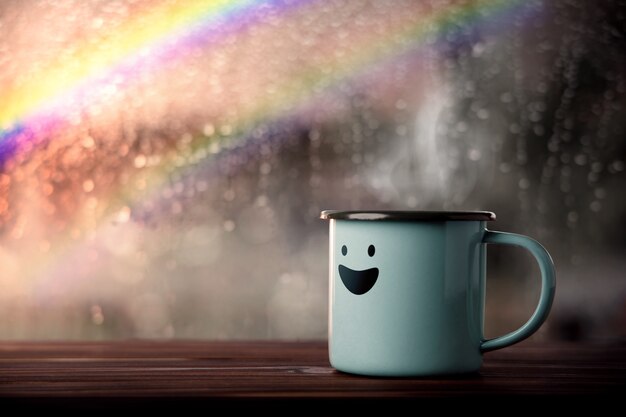 Felicità e mente positiva, concetto di salute mentale. Godersi il caffè con un cartone animato sorridente, pioggia sfocata con arcobaleno come vista esterna. Sorridi in un giorno di pioggia