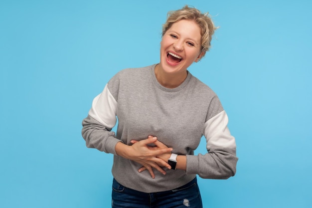 Felicità e risate. Donna estremamente eccitata con i capelli corti in felpa che tiene la pancia e ride ad alta voce, scoppiettando, divertita da uno scherzo divertente. tiro in studio indoor isolato su sfondo blu