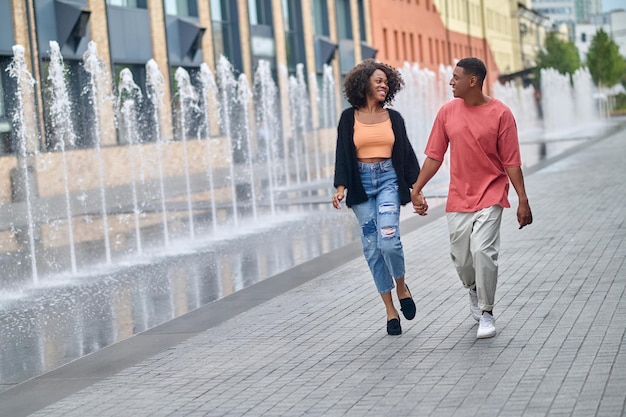 Felicità. Giovane uomo sorridente dalla carnagione scura e donna snella con i capelli ricci in abiti casual eleganti che si guardano tenendosi per mano camminando vicino alle fontane sulla strada della città