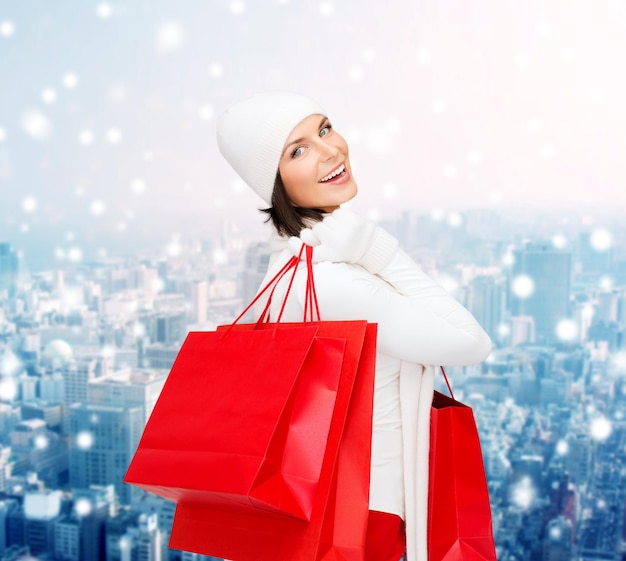 felicità, vacanze invernali, natale e concetto di persone - giovane donna sorridente con cappello bianco e guanti con borse della spesa rosse su sfondo di città innevata