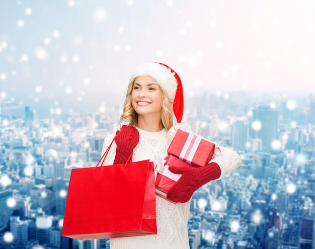 felicità, vacanze invernali, natale e concetto di persone - giovane donna sorridente con cappello da assistente di Babbo Natale con scatole regalo e borsa della spesa su sfondo città innevata