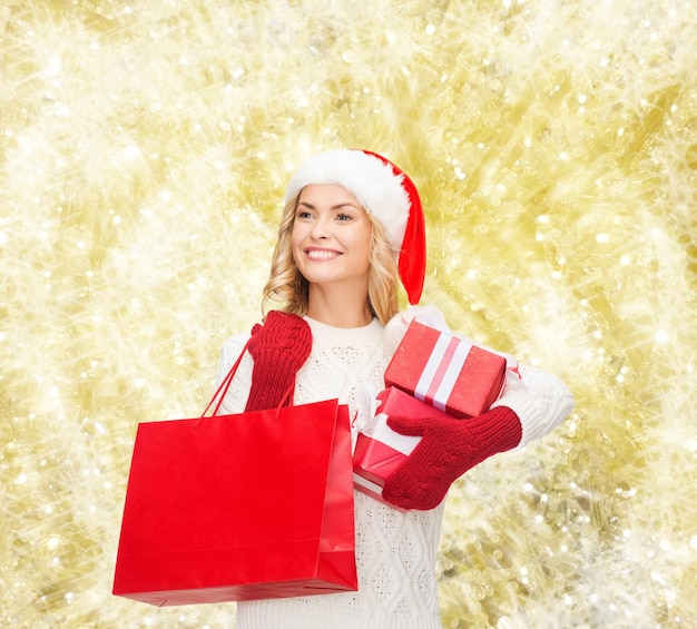 felicità, vacanze invernali, natale e concetto di persone - giovane donna sorridente con cappello da Babbo Natale con scatole regalo e borsa della spesa su sfondo a luci gialle