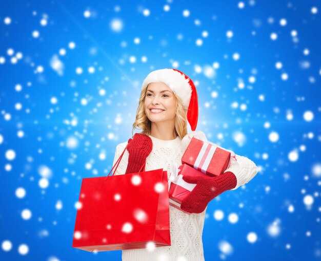 felicità, vacanze invernali, natale e concetto di persone - giovane donna sorridente con cappello da Babbo Natale con scatole regalo e borsa della spesa su sfondo blu nevicato