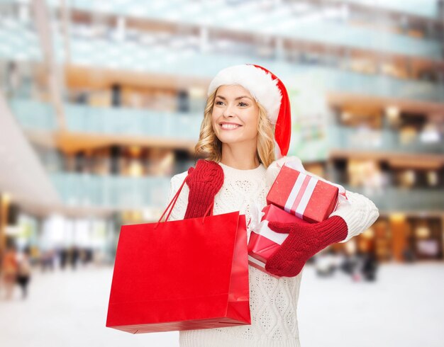 felicità, vacanze invernali, natale e concetto di persone - giovane donna sorridente con cappello da Babbo Natale con scatole regalo e borsa della spesa su sfondo del centro commerciale