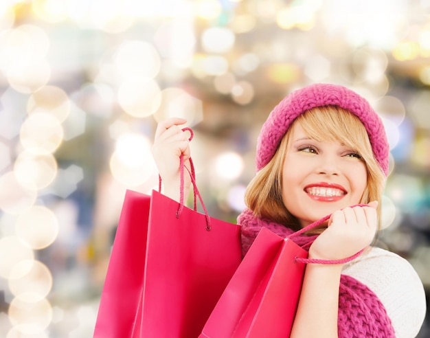 felicità, vacanze invernali, natale e concetto di persone - giovane donna sorridente in cappello e sciarpa con borse della spesa rosa su sfondo di luci