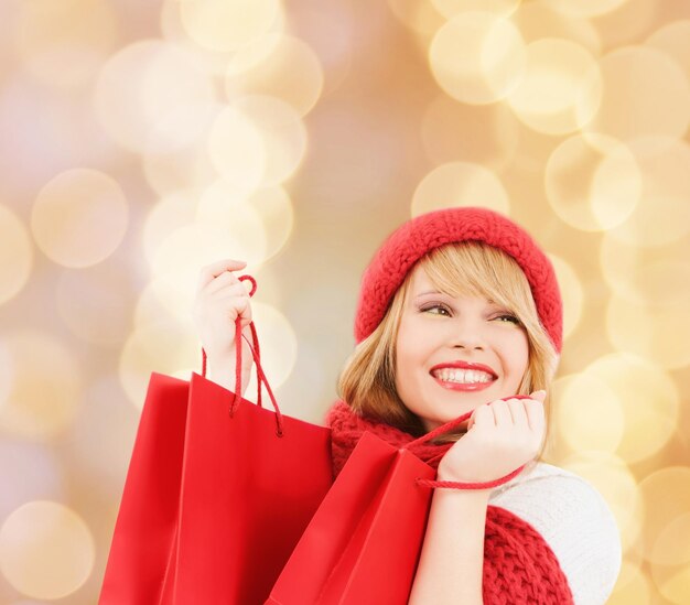 felicità, vacanze invernali, natale e concetto di persone - giovane donna sorridente in cappello e sciarpa con borse della spesa rosse su sfondo di luci beige