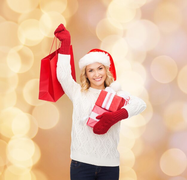 felicità, vacanze invernali, natale e concetto di persone - giovane donna sorridente in cappello santa helper con regali e shopping bag su sfondo di luci beige