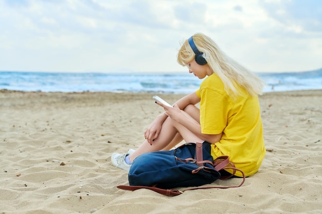 Femmina adolescente con zaino in cuffia utilizzando lo smartphone sulla spiaggia sabbiosa