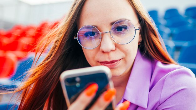 Femmina adulta concentrata con lunghi capelli castani in camicetta e occhiali seduta sul sedile della tribuna blu e messaggistica sullo smartphone