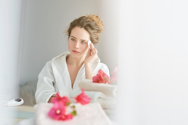 Femmina affascinante in accappatoio che asciuga la faccia con un cotone mentre guarda lo specchio vicino al tavolo con gli asciugamani durante la routine mattutina