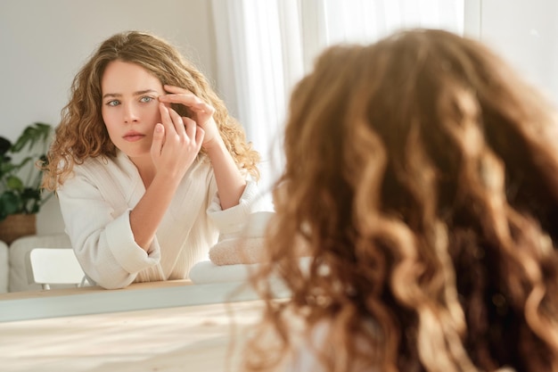 Femmina affascinante in accappatoio che tocca la faccia e guarda le rughe vicino agli occhi nello specchio mentre è seduta a tavola durante la routine mattutina a casa