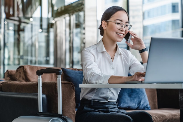 Femmina asiatica che si siede all'aeroporto prima del viaggio con una conversazione telefonica con il cliente e sorridente