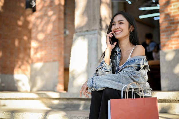 Femmina asiatica con le sue borse della spesa che parlano al telefono mentre si siede sulle scale della strada