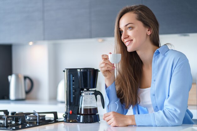 Femmina attraente felice che beve caffè aromatico fresco caldo dopo la preparazione del caffè facendo uso della macchinetta del caffè nella cucina a casa. Frullatore per caffè ed elettrodomestici da cucina per bevande calde