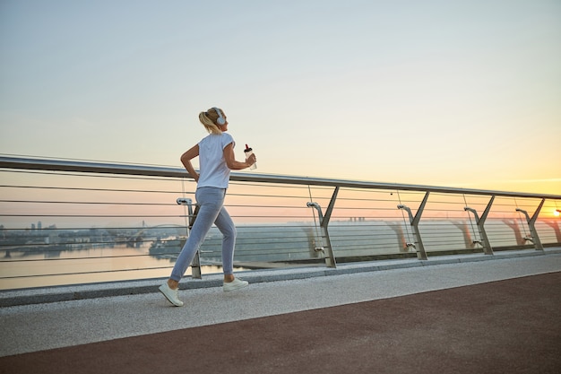 Femmina bionda atletica in cuffia e con una bottiglia d'acqua che fa jogging attraverso la passerella