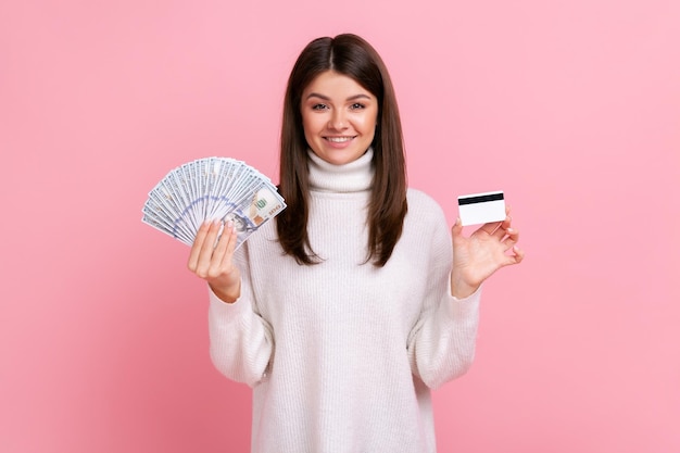 Femmina bruna positiva che tiene carta di credito e grande fan delle banconote in dollari, operazioni bancarie, guadagni, indossa un maglione bianco in stile casual. Studio indoor girato isolato su sfondo rosa.